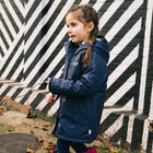 A girl wearing a navy Puddleflex Waterproof Fleece Lined Jacket stands against a striking black and white striped wall.